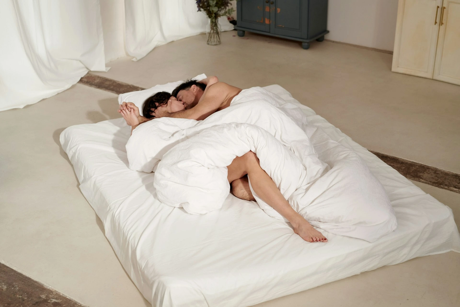 Two people embracing and kissing under a white duvet on a floor mattress